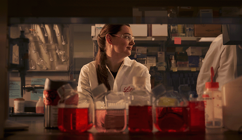 Scienziata in laboratorio che osserva il suo lavoro, circondata da strumenti e contenitori con liquidi rossi.